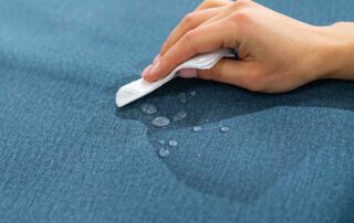 "A hand wiping water off a blue fabric surface, demonstrating the importance of maintaining and replacing awning fabric when needed. This image highlights the process of awning fabric care and the possibility of replacement for longevity and improved appearance.