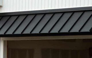A sleek black metal awning extending over the entrance of a white building exterior, providing both shade and architectural appeal. The contrast between the dark awning and the bright façade highlights durability and style, making it a great example of what to look for in a metal awning.